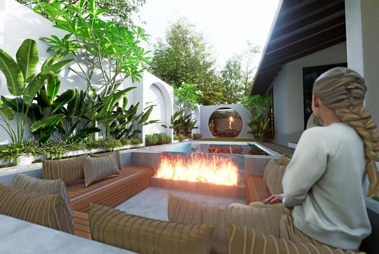 A woman with braided hair sits on a cushioned bench near a modern outdoor fire pit, overlooking a small pool surrounded by lush greenery and white walls in a contemporary courtyard.